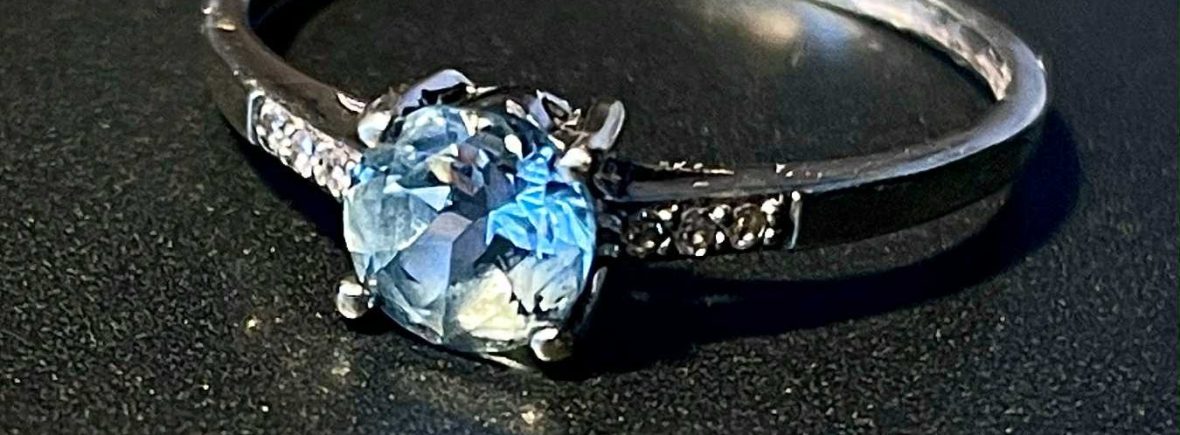 Bague en or blanc 375 millièmes avec une aigue-marine ronde centrale entourée de six diamants sur un fond sombre.