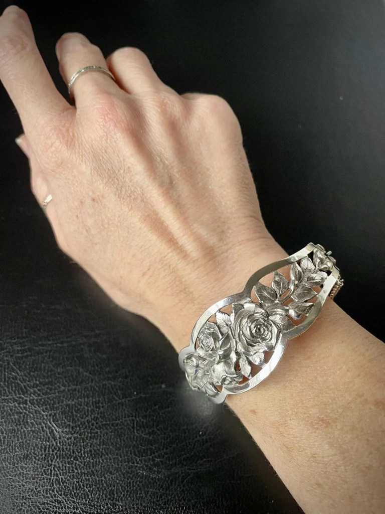 Vue portée d'un bracelet jonc d'orfèvrerie à décor floral. La mise en situation permet d'apprécier la largeur du bandeau ciselé de roses et le travail de découpe du métal. Fond noir texturé, lumière naturelle mettant en valeur la patine du bijou.