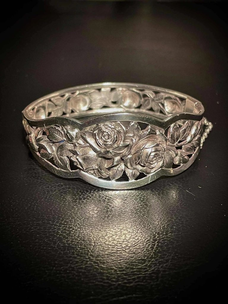Photographie de trois-quarts d'un bracelet jonc articulé en métal argenté, posé sur un support noir. Le décor central présente des roses sculptées en haut-relief et des feuilles ciselées. La chaînette de sécurité à mailles fines est visible sur le côté droit.