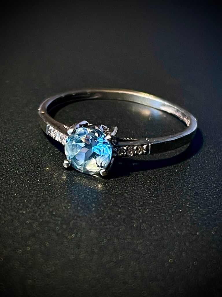 Bague en or blanc 375 millièmes avec une aigue-marine ronde centrale entourée de six diamants sur un fond sombre.