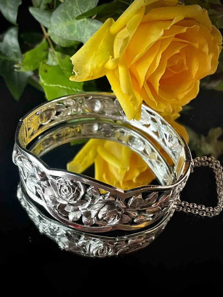Bracelet jonc en argent ciselé avec un décor de roses en relief, posé sur un miroir à côté d'une rose jaune